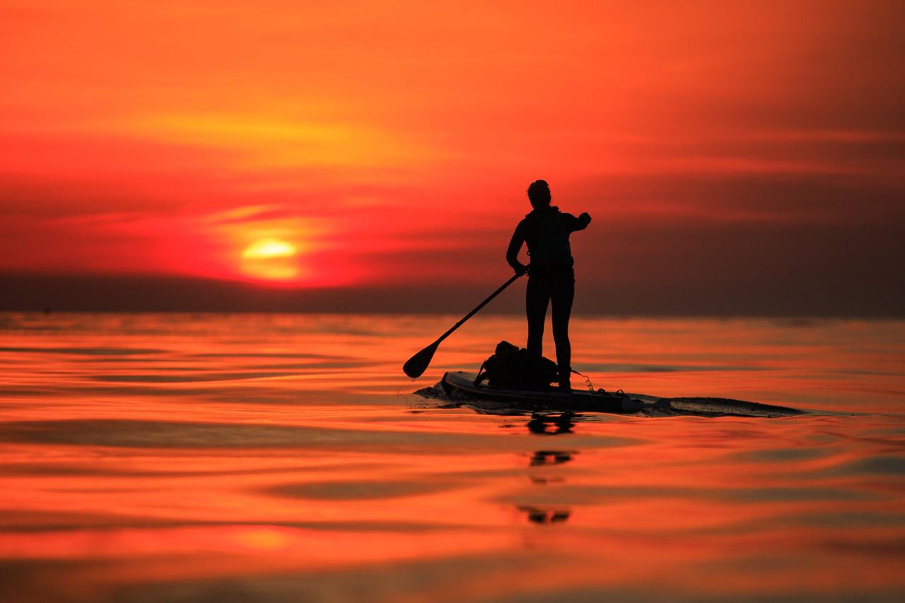 A woman is shown as a silhouette on a stand up paddle board on the water, with the sun setting against a very orange sky, reflecting this colour back onto the water.