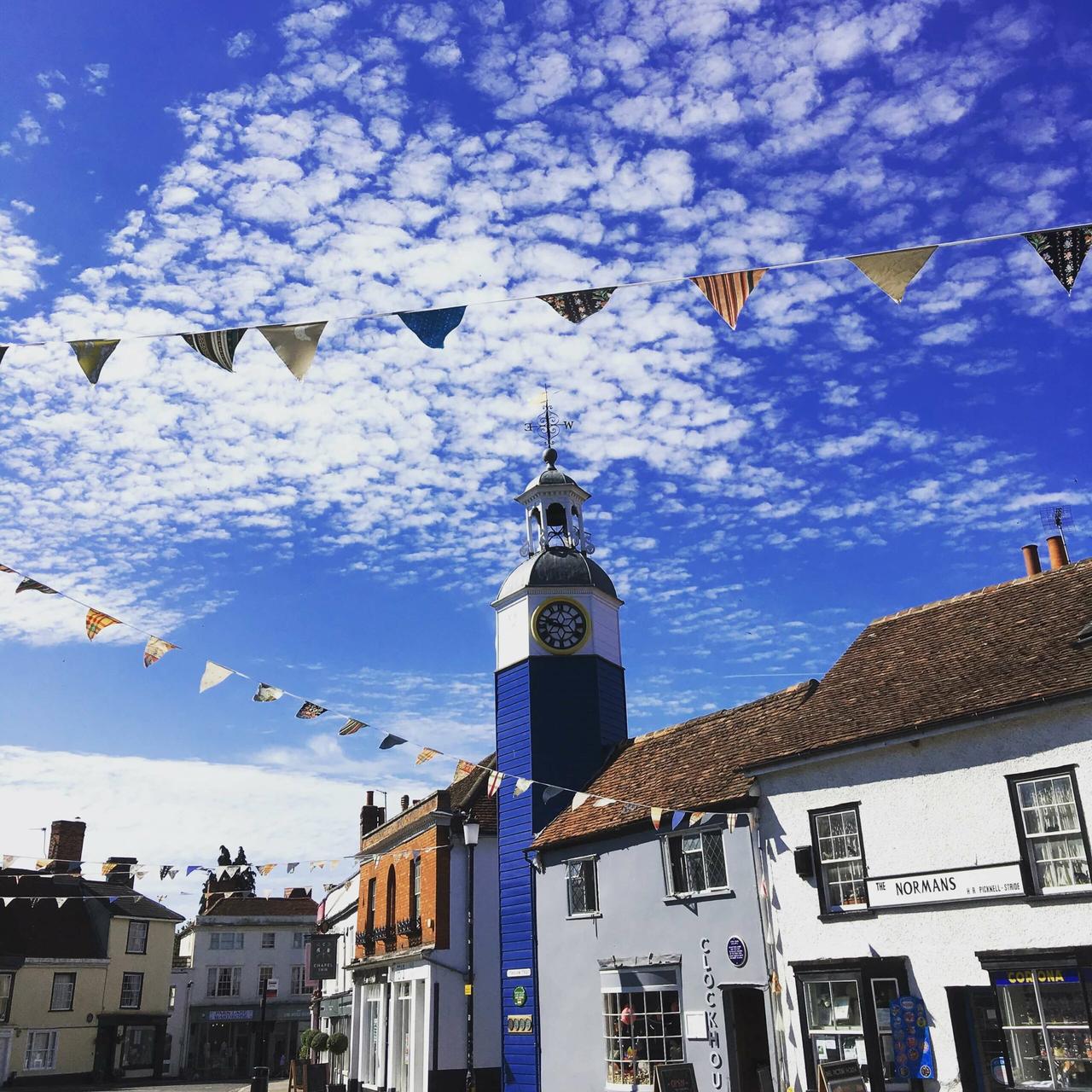 A vibrant town square with a blue clock tower in the centre, surrounded by historic buildings. Colourful bunting hangs across the scene against a bright blue sky filled with fluffy white clouds. The buildings include a white structure with a sign reading "The Normans" and a shop with a display window.