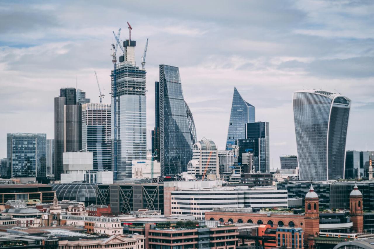 A panoramic view of a modern city skyline featuring various skyscrapers. In the foreground, there are red-brick buildings with arched windows and a clock tower. Prominent structures include a partially constructed tall building with cranes, a glass building with a distinctive curvy shape, and others with angular designs. The skyline is set against a cloudy sky, with a mix of glass and metal facades reflecting light. The scene captures the juxtaposition of historical architecture and contemporary designs.