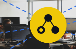 In a bright office space, two people are seated at desks working on computers. One person, with dark hair and wearing a striped top, is visible in the foreground, while another person is partially obscured behind a computer monitor. The scene is lively, with large windows allowing natural light to filter in. In the centre of the image, a large yellow circle features a black graphic resembling a network or connection, with three nodes and lines connecting them. Blue lines radiate from the circle, adding a technical feel.