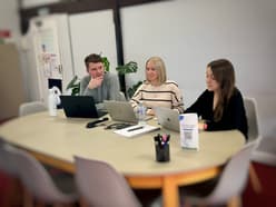 Meeting to discuss website design and development with three people: two woman and one man, all with laptops sitting at a boardroom table. On the table are two water bottles, a notepad, pen, cables, pot of pens and a standing advert with a QR code on it. In the background is a plant and there are beams on the wall. There is a door on the far top left, exiting out of the office.