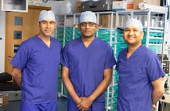 Three doctors in scrubs look directly at the camera, either smiling or appearing thoughtful. They all wear scrub caps on their heads.