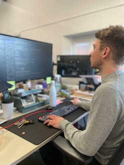 A person sits at a desk, focused on two computer monitors displaying lines of code. They are wearing a light grey sweater and have short hair. The desk is cluttered with various items, including a white water bottle, a disposable coffee cup, and a bowl with food. A black mouse pad holds a mechanical keyboard, with a red cable connected to it. Keys and a remote control are scattered on the desk. The workspace is well-lit, with natural light coming from a window in the background.