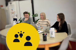 Meeting to discuss ideas between three team members at a table with open laptops and plants in the background. On the bottom left is a large yellow circle containing two black silhouette icons and a black lit lightbulb next to one of them.