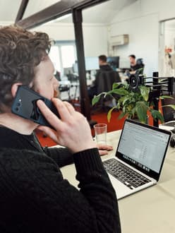 A man with curly hair and a beard is sitting at a desk in an office environment, talking on a smartphone held to their ear. They are wearing a black knitted sweater and are focused on a laptop in front of them, which displays a webpage with data and charts. A glass of water is placed on the desk next to the laptop, and a green potted plant is visible to the right. In the background, two other individuals can be seen working at separate desks, with computers and office equipment