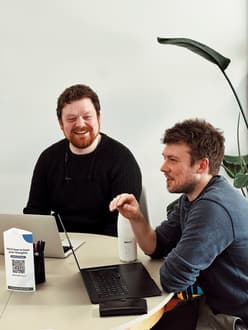 Two men are seated at a round table in a bright, modern office space. The man on the left has curly brown hair and a beard, wearing a black knitted sweater, and is smiling. The man on the right has short, tousled hair and is dressed in a grey long-sleeve shirt, gesturing with his hand as he speaks. Both have laptops open in front of them. A black pen holder with colourful pens is on the table, along with a white water bottle