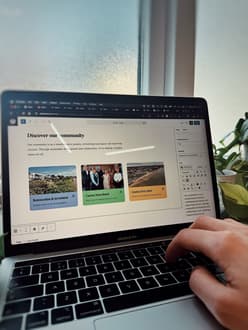 A person is typing on a laptop keyboard, with a focus on the screen displaying a webpage titled "Discover our community." The webpage features three coloured sections: the first section, green, titled "Regeneration & investment," includes an image of a coastal landscape; the second section, blue, titled "Clacton Town Board," shows a group of people in formal attire; the third section, orange, titled "Clacton Town vision," features an aerial view of a town.