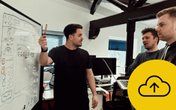 A group of three men are engaged in a discussion in a modern office space. One man, standing in front of a whiteboard, is holding a red marker and pointing at a diagram on the board. The whiteboard features various sketches, including circles and boxes, likely representing a user interface or design elements. Two other men, positioned to the right of the speaker, are observing intently. There is a yellow circle on the bottom right of the image with a cloud icon contain an upwards pointing arrow.