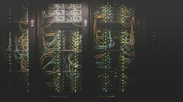 A close-up image of a server rack, featuring multiple rows of servers behind a mesh panel. The servers are connected with numerous cables, primarily in blue and orange, which weave in and out of the rack. Small green lights on the servers indicate activity, with some areas having brighter lights than others. The overall setting appears dark, highlighting the illuminated cables and lights, creating a high-tech atmosphere.