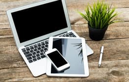 A wooden desk with a small grass style potted plant to the right, a pen and then on the left there is an Apple laptop and on top of it is an Apple tablet and an Apple phone.
