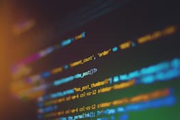 Close-up image of computer code on a dark screen, featuring lines of programming syntax in various colours, including blue, yellow, and white. The code appears to be related to a web application, with functions and parameters visible. A soft orange glow is present on the left side of the image.
