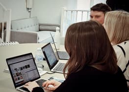A group of three people is seated around a round table, each using a laptop. The focus is on a woman with long brown hair, typing on her laptop, which displays a webpage with images. Next to her, a water bottle and a notebook are visible. In the background, a light grey sofa is positioned against a wall, and a man with short dark hair is partially visible, looking towards the group. Another woman, with short blonde hair, is also present, facing the table.