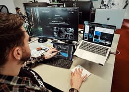 A person with short hair and a beard is seated at a desk in a modern office environment. They are wearing a black shirt and a plaid shirt over it. The individual is using a stylus on a tablet, which displays a design interface. To the left, there is a large monitor showing a dark-themed gallery webpage featuring natural imagery. On the right, a laptop is open, also displaying a gallery with various images. The desk is cluttered with a few items, including a brown case