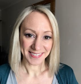A smiling woman with shoulder-length blonde hair is facing the camera. She has a light complexion and is wearing subtle makeup, including mascara and a natural lip colour. She is dressed in a grey top with a teal cardigan over it. The background is softly blurred, suggesting an indoor setting with neutral walls. The overall mood of the image is friendly and approachable.