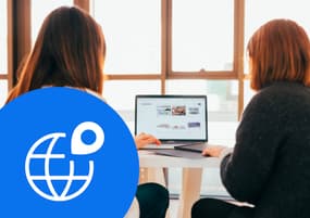 Two woman looking at a laptop screen. On the bottom right is a large circle that is cropped by the edges of the image. It is blue and inside is a white icon of a globe used for the internet with a location pin on it.