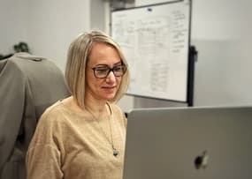 A woman with short blonde hair and glasses is seated at a desk, looking at a laptop screen. She is wearing a beige sweater and a necklace. In the background, there is a whiteboard with handwritten notes and diagrams. A grey coat is draped over the back of her chair.