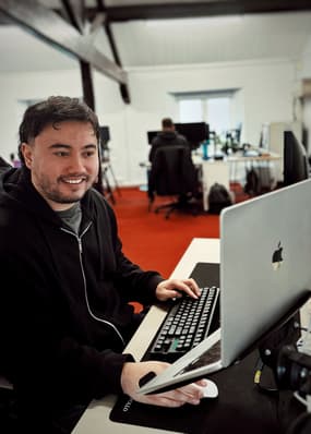A man is smiling whilst working in an office at an open laptop