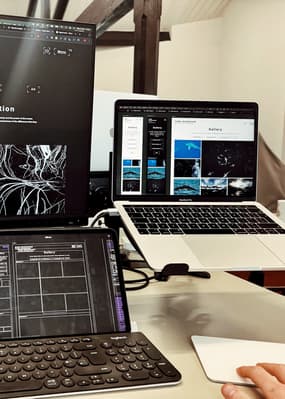 A workspace featuring two laptops and a tablet on a desk. The left laptop displays a dark interface with a black and white abstract image, while the right laptop shows a gallery layout with images of ocean life and a heading that reads "Gabe Hearnshew." Below the right laptop, a tablet shows a wireframe design for a gallery, with empty rectangular sections outlined in purple. A hand is positioned on a white trackpad, indicating active engagement with the devices. The background includes wooden beams.