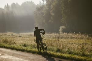 A cyclist in a black outfit stands on a quiet road, facing away from the camera. They are holding a camera in their right hand and adjusting their bike with their left hand. The background features a misty landscape with tall grass and scattered wildflowers, illuminated by soft morning light. Dense trees line the horizon, creating a serene atmosphere. The road is bordered by green grass, and gentle sunlight filters through the mist, enhancing the tranquil scene.