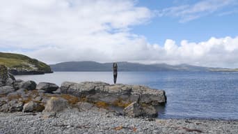 A person stands on a large rock by the water, facing away from the camera. They are wearing a dark jacket and black trousers, with arms raised above their head. The scene features a calm sea reflecting the sky, surrounded by green hills and distant mountains under a partly cloudy sky. The foreground includes smooth grey pebbles and patches of seaweed, while the background showcases rolling hills and a few clouds scattered across the blue sky.