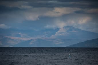 A woman is shown as a small speck on the sea, stand up paddle boarding. There are huge mountains in Scotland behind her and the sky is grey, cloudy and somewhat ominous. The composition of the photo shows how incredible the landscape is and how far the woman is from the shore.