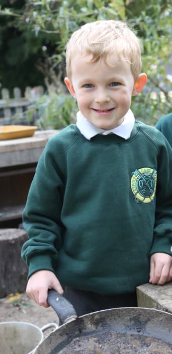 A young blonde boy smiling directly at the camera wearing a green school jumper with its logo on while in a school playground at the mud kitchen