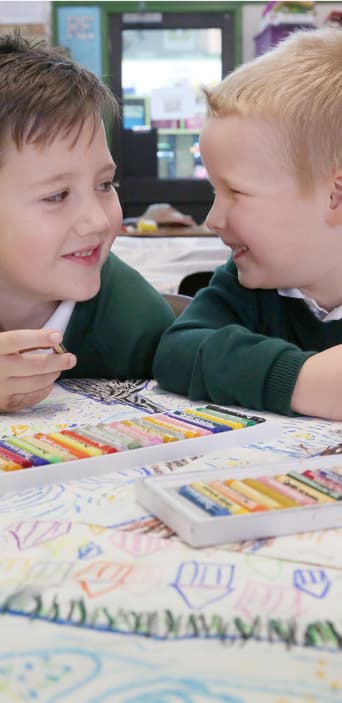 Two young boys looking at each other and smiling in front of colouring crayons in a classroom