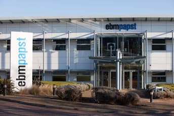 The image shows the exterior of an ebm-papst building. The main entrance features a glass structure with two doors, surrounded by grey metal pillars and a roof that has thin, slanted supports. The company's name, "ebm-papst," is prominently displayed in blue on the building's facade. To the left, a large white sign with the same name stands tall, with "ebm" in black and "papst" in blue. The landscape includes neatly trimmed bushes