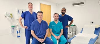 Four male doctors are in a consulting room. Two are standing with hands on hips, and the other two are seated with their hands clasped.