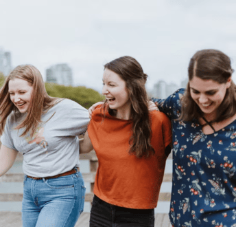 Three young woman have their arms around each other as they walk along, all smiling and laughing