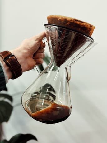 A hand holds a glass coffee maker with a brown filter on top, containing brewed coffee. The glass vessel has a round base and a narrow neck, with dark liquid visible at the bottom. The background is blurred, featuring a white wall and a green plant leaf, adding a natural touch to the scene. The person’s wrist is adorned with a brown leather watch, contrasting with a green sweater.