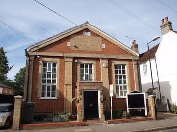 A red-brick building with a symmetrical façade, featuring large, multi-paned windows on the ground and first floors. The entrance, centred and slightly protruding, has a stone archway with two doors. Flanking the entrance are two planters with greenery. To the left, there is a green wheelie bin and a lamppost, while to the right, a white building is partially visible. The roof has a triangular shape, with a small chimney on the left side. The sky