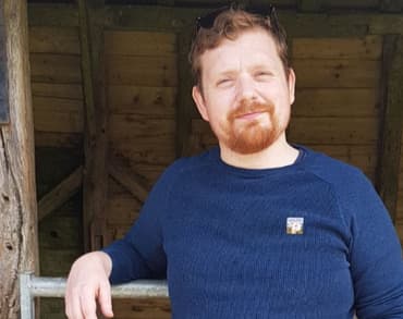 Hector Westcroft stares directly at the camera in a blue jumper with a wooden structure in the background, resting one arm on a metal gate