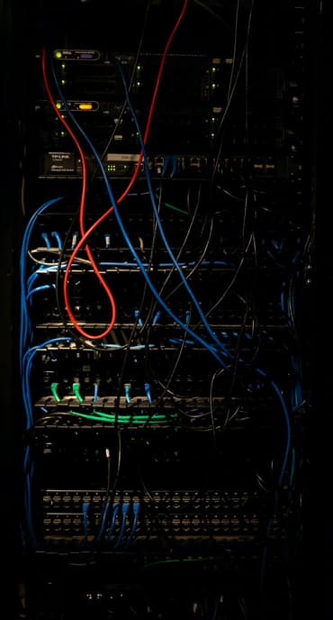 A close-up view of a server rack filled with various networking equipment and cables. The image shows multiple rows of devices with blinking lights, indicating activity. Numerous cables in different colours, including red, blue, and green, are tangled and connected to the equipment. The background is dark, highlighting the illuminated components.