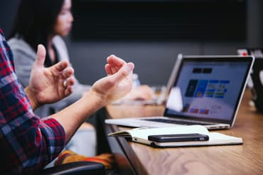 A man is gesturing while talking at a desk in front of a laptop with his phone next to him and a woman is blurred in the background.