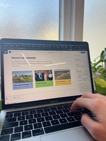 Wordpress CMS editor for a website that has the heading 'Discover our community' on the page. A person's hand is at the laptop, typing.