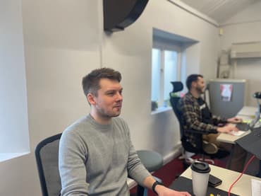 A man is smiling while looking at his computer. In the background a slightly blurred second man is laughing.