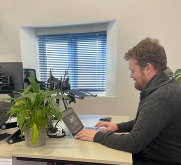 A man with red curly hair and a red beard sits at a desk with a laptop and a potted plant