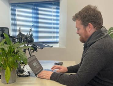 A man with red curly hair and a red beard sits at a desk with a laptop and a potted plant