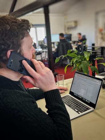 A person with curly hair is sitting at a desk, holding a smartphone to their ear while engaged in a phone call. They are wearing a dark green, knitted sweater. In front of them, a laptop is open, displaying a webpage with various data tables. A glass of water sits beside the laptop on a beige table. In the background, blurred figures of two colleagues are visible, working at their own desks with computers. A green potted plant is positioned near the laptop
