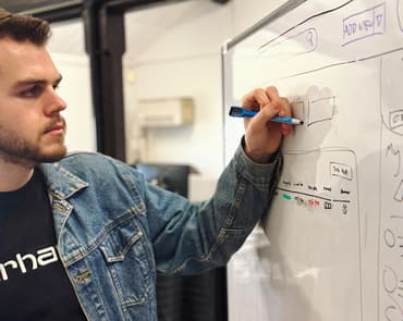 A young man with a beard, wearing a denim jacket and a black t-shirt, is drawing on a whiteboard. He is using a blue marker to outline boxes and diagrams. The whiteboard features various sketches, including interface elements and text, suggesting a brainstorming session or design work.