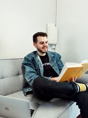 Man reading a user experience book on a sofa with an open laptop and smiling