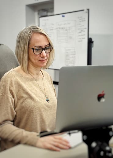 A woman with shoulder-length, straight blonde hair sits at a desk, focused on her laptop. She wears glasses and a light beige, oversized sweater, accessorised with a necklace featuring a turquoise pendant. Her right hand rests on a white keyboard, while the laptop screen displays a silver Apple logo. In the background, a whiteboard is visible, covered with handwritten notes and diagrams, suggesting a collaborative workspace. The overall atmosphere is professional and engaged.