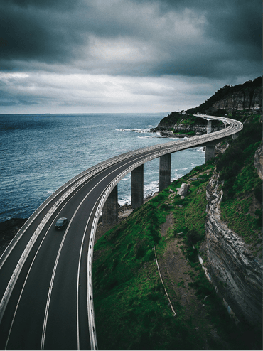 A winding road curves along a coastal cliff, supported by grey concrete pillars. The road is asphalt with white markings, and a dark car is driving towards the viewer. To the left, the deep blue ocean meets rocky shores, while the right side features lush green vegetation and steep cliffs. The sky is overcast with dark clouds, creating a dramatic atmosphere.