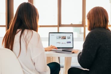 Two woman looking at a laptop screen with a window behind them
