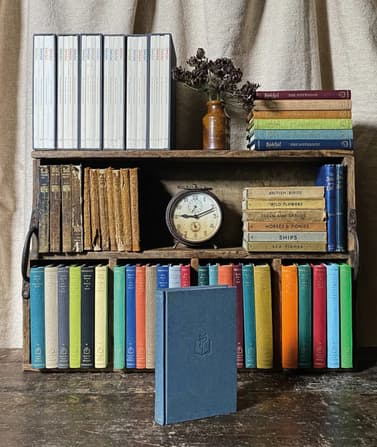 Three shelves as part of an old fashioned bookshelf containing a range of old books and new books from Slightly Foxed. There is also a clock in the middle shelf and a masonry jar of dried flowers on the top shelf. In front of these shelves is a single blue book standing upright.