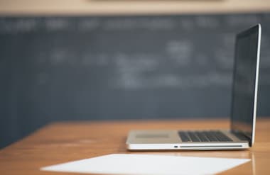 A silver laptop sits on a wooden table, angled to show its side. The laptop is open but the screen is dark, indicating it is not in use. In front of the laptop, there is a blank white sheet of paper placed flat on the table. In the background, a blackboard is visible with faint chalk markings, suggesting a classroom or study environment. The overall setting appears calm and conducive to learning or working.
