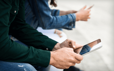 A close-up view of two people sitting on a surface, both engaged with their devices. The person on the left, wearing a dark green shirt, is holding a tablet with a reflective screen. The person on the right, wearing a blue denim jacket, is using a smartphone. Their hands are visible, with the focus on the tablet held by the individual on the left. The background is softly blurred, suggesting an outdoor setting.