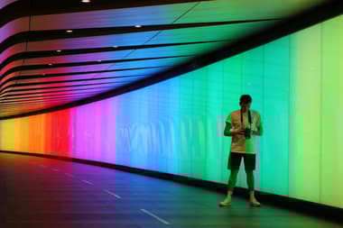 A man stands inside a tunnel that has panels running along it with different lights, showing rainbow colours. He is wearing a light t shirt, shorts, socks and trainers. He has a camera aroudn his neck and is looking at the camera screen.
