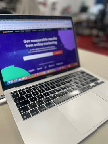 A close-up view of a laptop keyboard and screen, displaying a website for SEMRUSH. The screen features a dark purple background with green and purple shapes at the corners. The text reads "Get measurable results from online marketing" above a search bar. The laptop is placed on a light-coloured surface, with blurred office furniture in the background. The keys on the keyboard are black with white lettering, and the touchpad is silver.